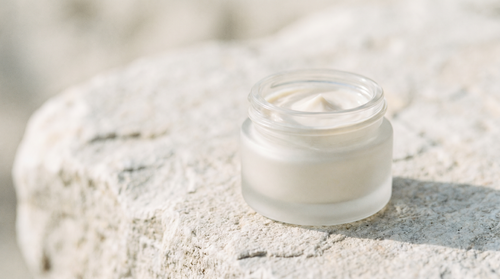 Small frosted glass jar of eye cream on pale stone in soft light.