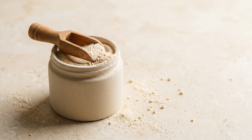 Matte jar of collagen powder with a wooden scoop and scattered powder.