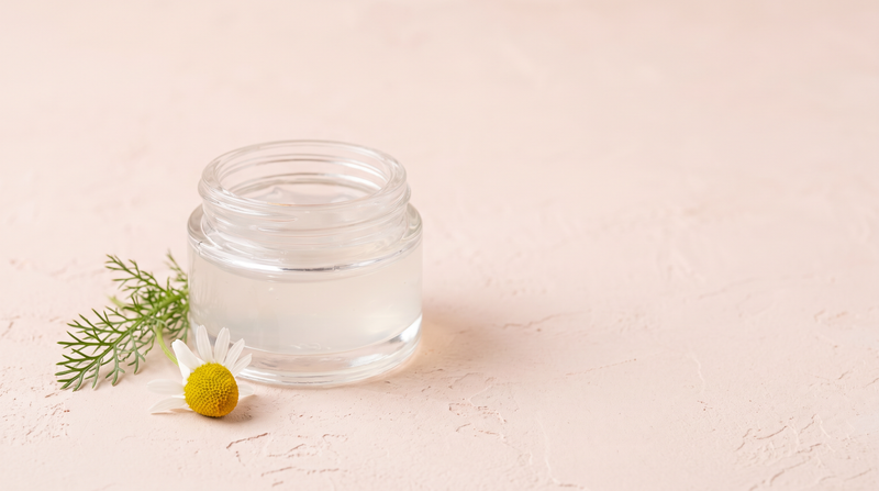 An open jar of clear gel on pale pink plaster beside a single chamomile flower.