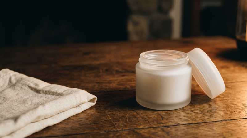 A white frosted glass cream jar with its lid ajar on warm oak in dusk-toned low light, folded ivory linen beside it