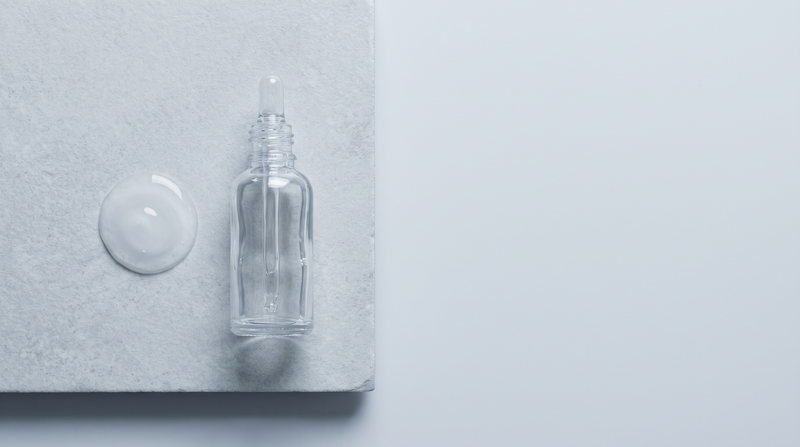 A clear glass dropper bottle on a pale stone slab beside a single bead of clear gel.