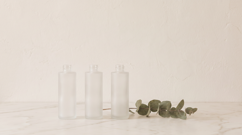 Three unlabeled frosted glass skincare vessels in a row on white marble with a eucalyptus stem.