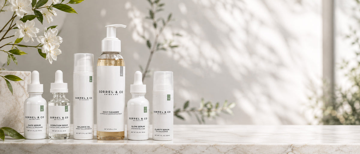 Six skincare bottles arranged on a light marble surface against a softly lit, sun-dappled white background with delicate green leaves and white flowers.